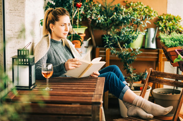 Young beautiful woman relaxing on cozy balcony, reading a book, wearing warm knitted pullover, glass of wine on wooden table