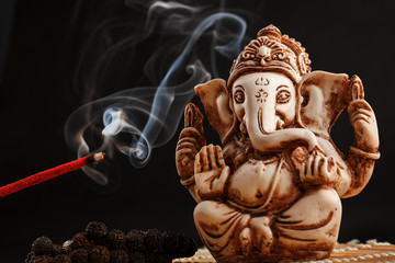 Hindu god Ganesh on a black background. Rudraksha statue and rosary on a wooden table with a red incense stick and incense smoke