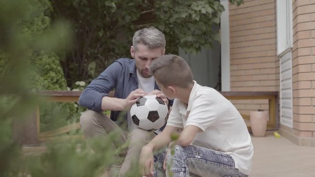 Portrait of father and his son sitting on the porch holding a deflated soccer ball in hands close-up. Family spending time together. Summertime leisure