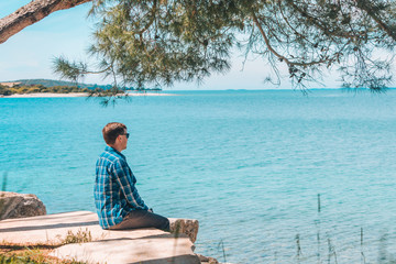 Fototapeta premium man sitting in the shadow of tree looking at blue azure water in sunny day