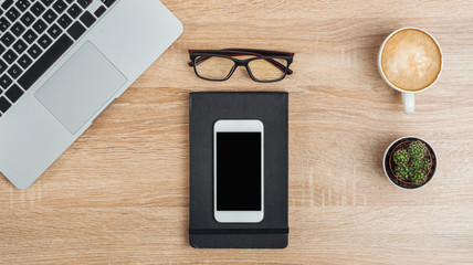 Smartphone with a dark screen on a notebook, laptop, glasses, cactus and a cup of coffee on the desktop. Top view. Mock up.