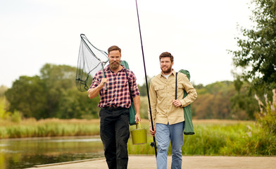 leisure and people concept - happy friends with fishing rods and scoop net walking along wooden pier at lake or river