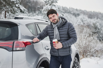 Portrait of car owner that standing near his automobile with cup of coffee in the hand at snowy winter day