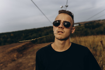 Young sexy man in black shirt and sunglasses standing outdoor. Dark key photo