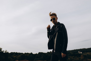 Young sexy man in leather jacket and sunglasses standing outdoor. Silhouette against grey sky