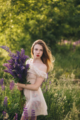 Beautiful girl in a dress with a bouquet of purple flowers stands in a field at sunset.