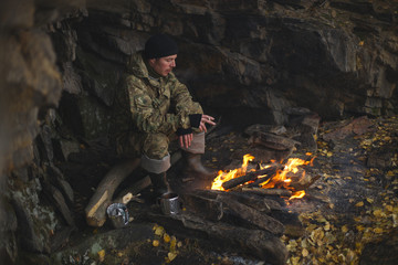 Traveler is warmed by the fire, hiding from the bad weather under an overhanging rock on a cold autumn day. Survival in the wild.