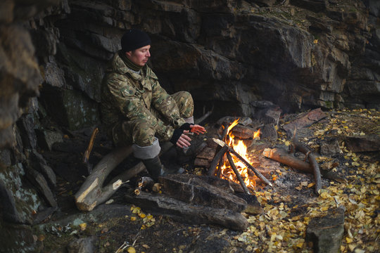 Traveler Is Warmed By The Fire, Hiding From The Bad Weather Under An Overhanging Rock On A Cold Autumn Day. Survival In The Wild.