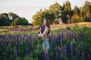 Beautiful girl in a dress with a bouquet of purple flowers stands in a field at sunset.