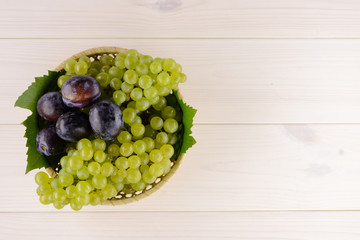 A bowl of tasty, ripe summer fruits on the white wooden table. In the bowl plums and white grapes. Top view, copy space