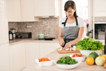 Beautiful smiling dedicated Caucasian brunette in apron standing in kitchen and chopping mushrooms. On table are lots of vegetables. Cooking at home concept.