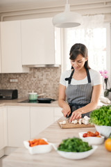 Beautiful smiling dedicated Caucasian brunette in apron standing in kitchen and chopping mushrooms. On table are lots of vegetables. Cooking at home concept.