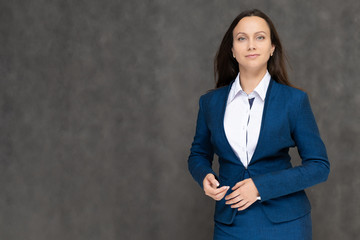 Portrait to the waist a young pretty brunette manager woman of 30 years in a business blue suit with beautiful dark hair. It is standing on a gray background, talking, showing hands, with emotions
