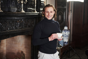 Young beautiful man near the luxury fireplace with white cup of tea