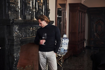 Looks at flames. Young guy with glass of wine in hands stands near the black fireplace
