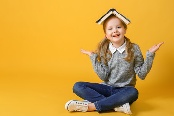 Cheerful little student girl is sitting with a book on her head and looking into the camera. The...