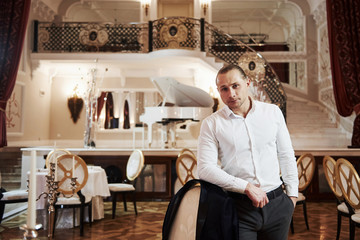 Portrait of guy in official wear stands in beautiful restaurant in vintage style with piano on the stage