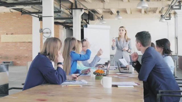 A female team leader in a business suit leads a presentation near a flipchart and describes business processes. Employees sit at a large table. Office life. Coworking
