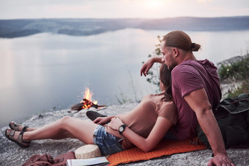 Hugging couple with backpack sitting near the fire on top of mountain enjoying view coast a river or lake. Traveling along mountains and coast, freedom and active lifestyle concept