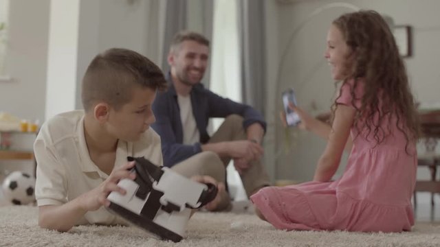 The boy and girl laying on the floor on fluffy carpet, the brother using virtual reality glasses, sister taking photo of father who sitting on the background of children. Happy friendly family at home