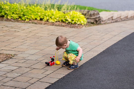 Side View Of Cute Fair Toddler Girl In Summer Clothes Crouched Holding The Rope Of A Pull Toy In A Driveway