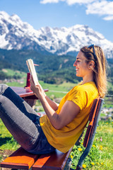 Young beautiful and happy girl reading a book on a bench, mountain range and greenery in the background.