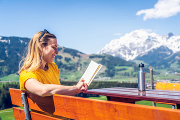 Young beautiful and happy girl reading a book on a bench, mountain range and greenery in the...