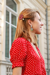 Photosession model with decorations of epoxy resin. Bijouterie. Red dress in peas. Straw hat. Stylish images. Fashion. Summer Trends 2019. Old architecture in the background.