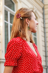 Photosession model with decorations of epoxy resin. Bijouterie. Red dress in peas. Straw hat. Stylish images. Fashion. Summer Trends 2019. Old architecture in the background.