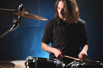 Portrait of a long-haired drummer with chopsticks in his hands sitting behind a drum set. Low key....
