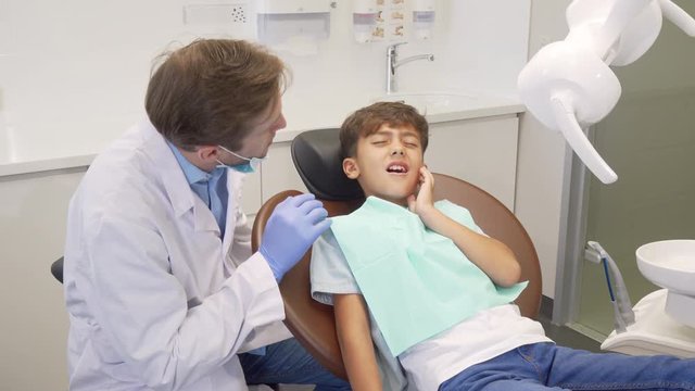 Little boy having toothache, sitting in dental chair during teeth examination. Professional dentist talking to his young patient with toothache. Pain, dental problems concept