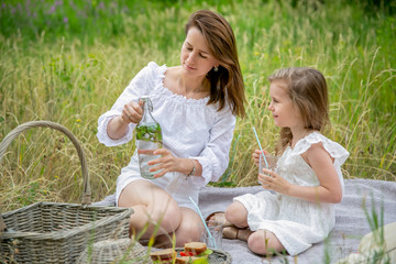 Beautiful young mother and her little daughter in white dress having fun in a picnic. They are sitting on a plaid and mom opens homemade lemonade. Maternal care and love. Horizontal photo