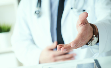 Male doctor making welcome gesture, politely inviting patient to sit down in medical office. Photo with depth of field. Just hands over the table.