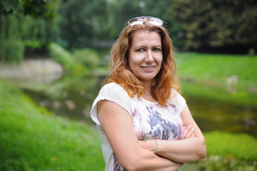 A good middle-aged woman is resting near the lake in the summer and smiles. The woman is looking forward to her husband to relax with him on the nature