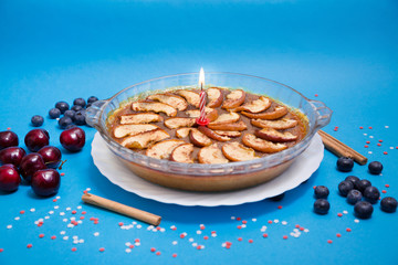 Tarta de manzana con canela, adornada con vela roja y blanca festiva, sobre fondo azul decorado con estrellas blancas y rojas, arándanos y cerezas.