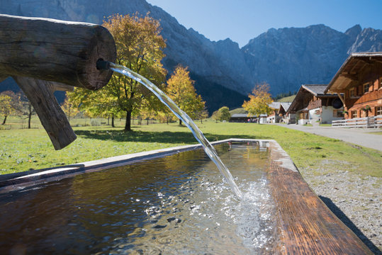 H&ouml;lzerne Tr&auml;nke, Brunnen mit Wasserstrahl, Almh&uuml;tten im Hintergrund