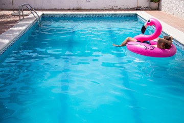 Fototapeta na wymiar Young man floating in a inflatable flamingo in a blue pool a sunny day of summer vacation 