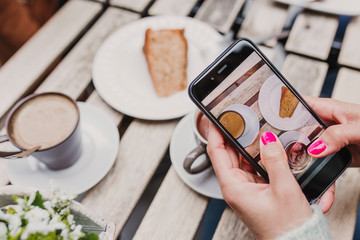Fototapeta na wymiar young unrecognizable woman taking a picture with mobile phone of her breakfast, a cup of coffee and a piece of cake. Breakfast concept