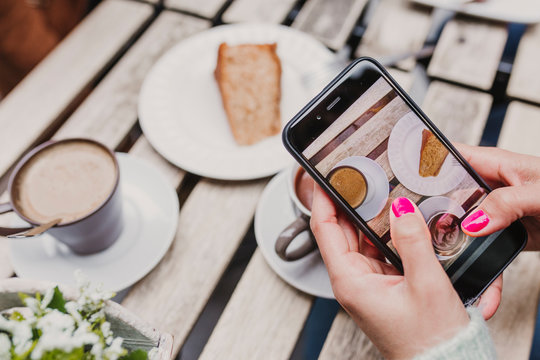young unrecognizable woman taking a picture with mobile phone of her breakfast, a cup of coffee and a piece of cake. Breakfast concept