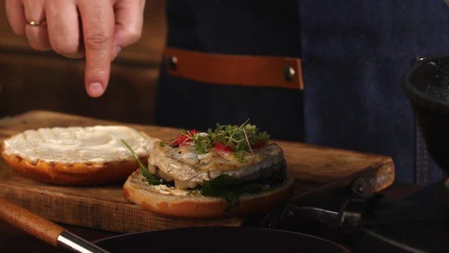 Close-up of male chef in blue apron adding greens on a fried hamburger bun. Stock footage. Preparing of delicious hamburgers