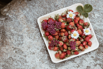 Fresh red strawberries in a wooden plate. Strawberry is a source of vitamin C. This berry is a natural aphrodisiac. Collage with wildflowers on the background of natural granite stone.