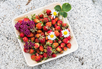 Fresh red strawberries in a wooden plate. Strawberry is a source of vitamin C. This berry is a natural aphrodisiac. Collage with wildflowers on the background of natural granite stone.