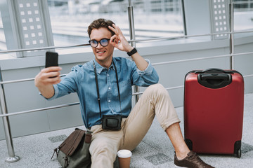 Happy man with camera using mobile phone and sitting in corridor in airport