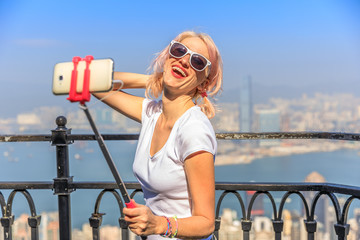 Tourist taking selfie stick picture with smart phone, enjoying view over Victoria Harbour from viewing platform on top of Peak Tower, Victoria Peak, Hong Kong, China