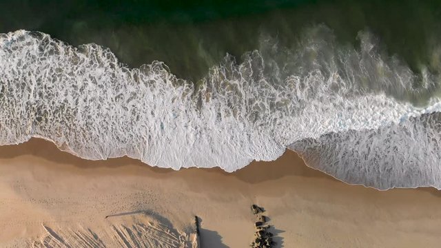 Aerial top down view of waves coming in on a pristine Leblon beach with the mouth of a drainage canal contrived of partly constructed and partly placed rocks starting from the city lake.