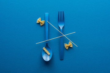 top view of spaghetti on blue plastic spoon upside down and fork near two kinds of pasta on blue...