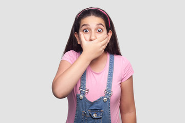 Portrait of shocked brunette young girl in pink t-shirt and blue denim overalls standing, covering her mouth and looking at camera with big eyes. indoor studio shot, isolated on light gray background.