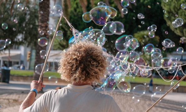 Street performer blowing soap bubbles outdoor. Young man entertains tourists on the street in summer. Funny caucasian man with blond curly hair makes big colorful soap bubbles.