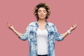 Portrait of calm young woman with curly hairstyle in casual blue shirt standing with closed eyes, raised arms, smile and doing yoga meditating. indoor studio shot, isolated on pink background.