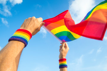 Hands with rainbow color wristbands waving LGBTQI gay pride flag in the wind against a vibrant sunny blue sky